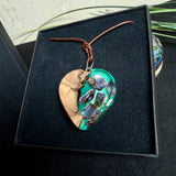 Herzförmiger Anhänger aus Olivenholz mit aquamarinfarbenem Harz und eingelegten Paua-Muschelstücken (Abalone), irisierender Blau-Grün-Schimmer. Der Anhänger hängt an einem braunen, verstellbaren Lederband mit Schiebeknoten; Aufhängung aus Edelstahl. Produktfoto vor maritimer Deko/Abalone-Schale.