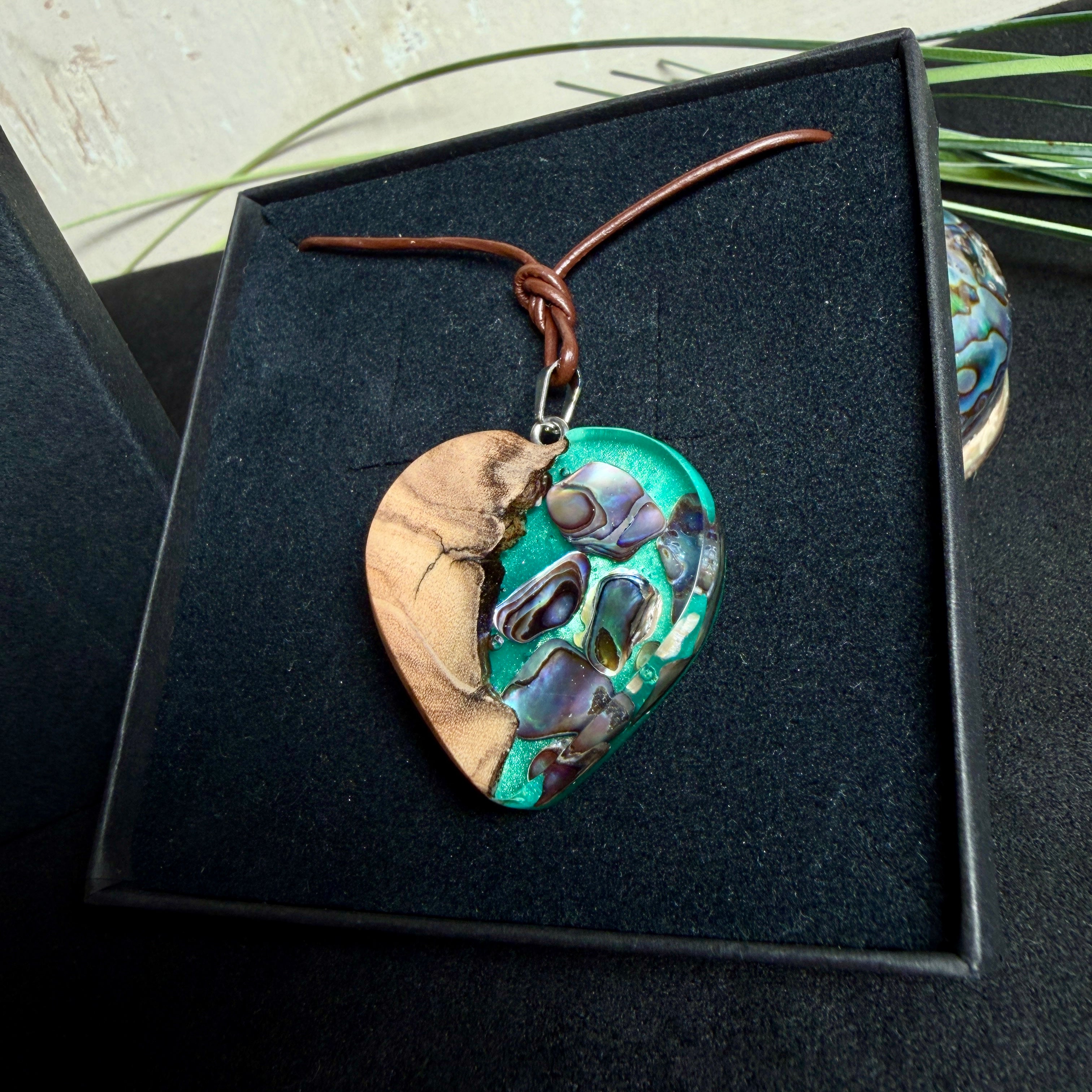 Herzförmiger Anhänger aus Olivenholz mit aquamarinfarbenem Harz und eingelegten Paua-Muschelstücken (Abalone), irisierender Blau-Grün-Schimmer. Der Anhänger hängt an einem braunen, verstellbaren Lederband mit Schiebeknoten; Aufhängung aus Edelstahl. Produktfoto vor maritimer Deko/Abalone-Schale.