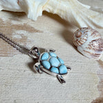Feine Kette aus rhodiniertem 925 Silber mit Wasserschildkröten-Anhänger. Der ca. 25 x 18 mm große Anhänger zeigt eine Schildkröte, deren Panzer aus mehreren hellblauen Larimar-Inlays besteht. Seltener, kostbarer Larimar mit typischen wolkigen Strukturen, an einer ca. 45 cm langen, 1 mm starken, auf Platin rhodinierten Silberkette. Fotografiert in schwarzer Schmuckbox und vor maritimer Dekoration mit Muscheln.