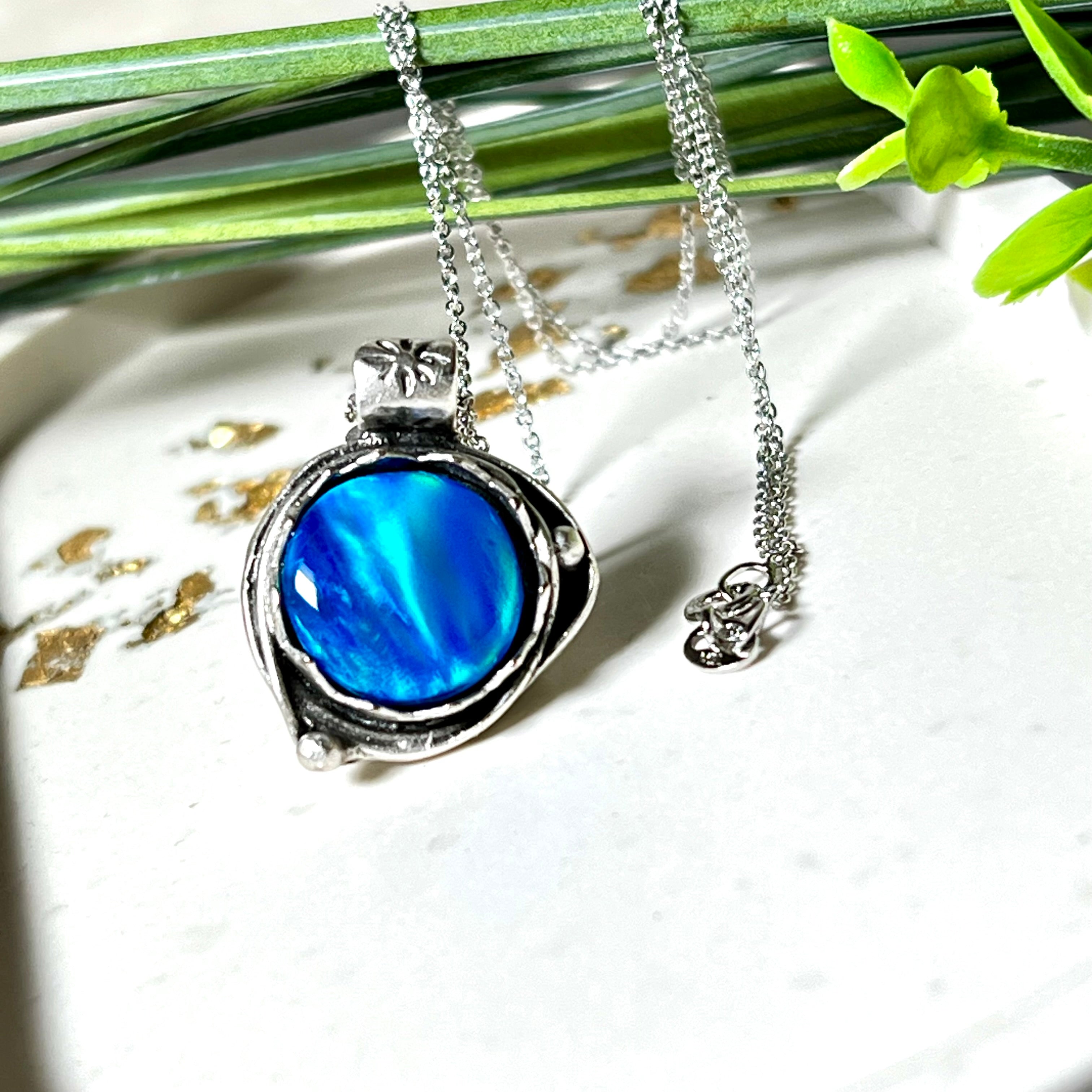 Handgemachter Anhänger mit rundem Cabochon aus Epoxidharz und schimmerndem synthetischem Opal. Die Farben changieren je nach Lichteinfall in Blau, Türkis und Grün. Der Opal sitzt in einer antik versilberten Fassung aus Messing (blei- & nickelfrei). Geliefert mit einer rhodinierten 925 Silberkette (45 cm). Jedes Stück ein Einzelstück – perfekt als stilvolles Geschenk oder persönlicher Glücksbringer.