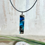 Rechteckiger Stabanhänger aus Maserholz und glasklarem Epoxidharz in tiefen Blau- und Türkistönen mit organischer Lagunen/Riff-Optik. Hochglanzpoliert, ca. 4,6 × 1,2 cm. Der Anhänger hängt an einer schwarzen Lederkette mit Edelstahl-Karabiner und 5-cm-Verlängerung (45 + 5 cm); die Anhängerschlaufe ist ebenfalls aus Edelstahl. Handgefertigtes Unikat, Lieferung in edler Schmuckbox – maritimer Look.