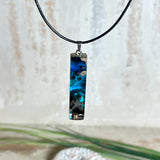 Rechteckiger Stabanhänger aus Maserholz und glasklarem Epoxidharz in tiefen Blau- und Türkistönen mit organischer Lagunen/Riff-Optik. Hochglanzpoliert, ca. 4,6 × 1,2 cm. Der Anhänger hängt an einer schwarzen Lederkette mit Edelstahl-Karabiner und 5-cm-Verlängerung (45 + 5 cm); die Anhängerschlaufe ist ebenfalls aus Edelstahl. Handgefertigtes Unikat, Lieferung in edler Schmuckbox – maritimer Look.