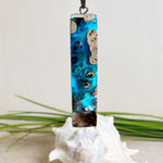 Rechteckiger Stabanhänger aus Maserholz und glasklarem Epoxidharz in tiefen Blau- und Türkistönen mit organischer Lagunen/Riff-Optik. Hochglanzpoliert, ca. 5,3 × 1,2 cm. Der Anhänger hängt an einer schwarzen Lederkette mitk Edelstahl-Karabiner und 5-cm-Verlängerung (45 + 5 cm); die Anhängerschlaufe ist ebenfalls aus Edelstahl. Handgefertigtes Unikat, Lieferung in edler Schmuckbox – maritimer Look.