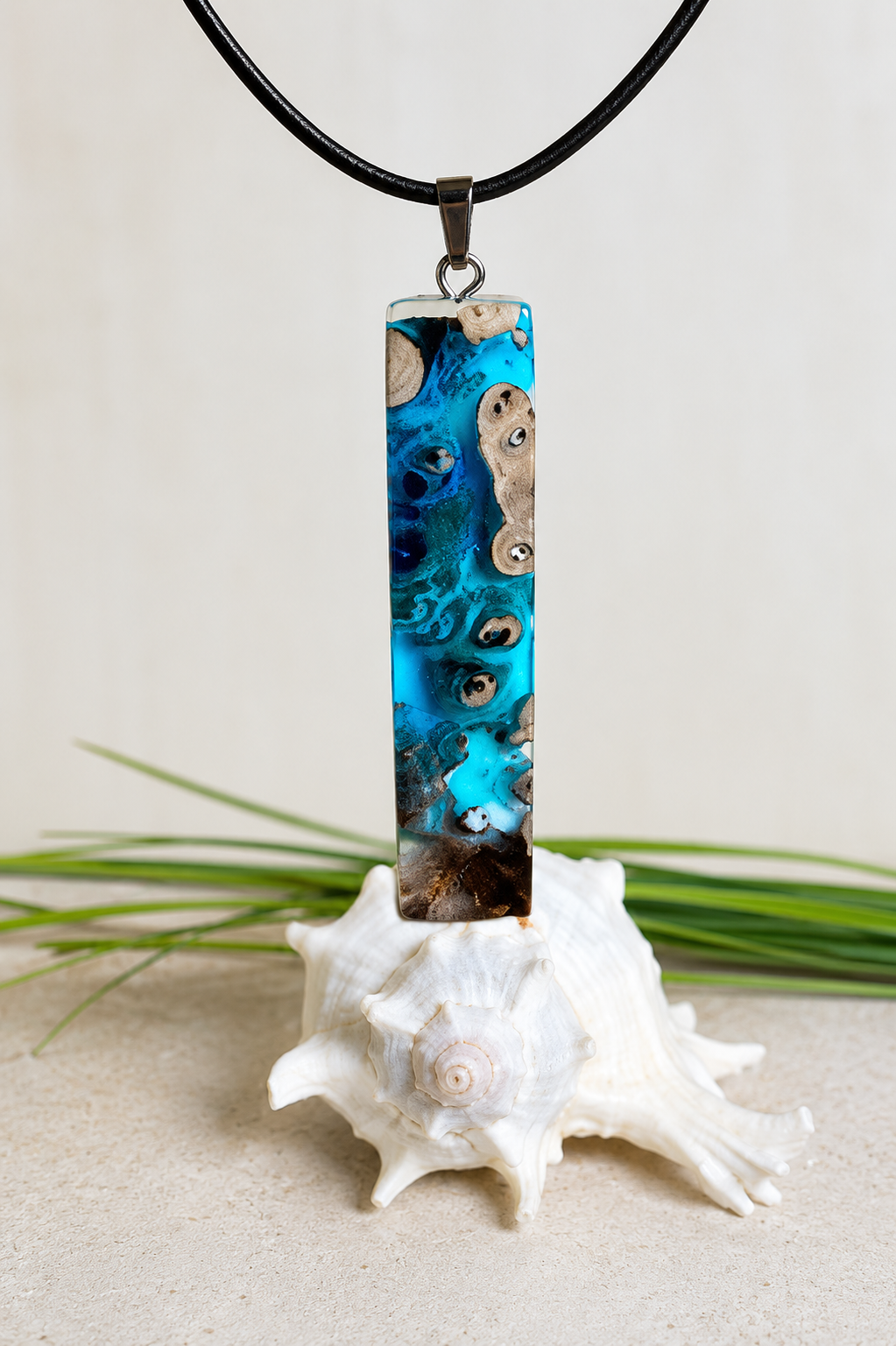 Rechteckiger Stabanhänger aus Maserholz und glasklarem Epoxidharz in tiefen Blau- und Türkistönen mit organischer Lagunen/Riff-Optik. Hochglanzpoliert, ca. 5,3 × 1,2 cm. Der Anhänger hängt an einer schwarzen Lederkette mitk Edelstahl-Karabiner und 5-cm-Verlängerung (45 + 5 cm); die Anhängerschlaufe ist ebenfalls aus Edelstahl. Handgefertigtes Unikat, Lieferung in edler Schmuckbox – maritimer Look.