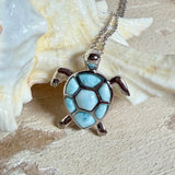 Feine Kette aus rhodiniertem 925 Silber mit Wasserschildkröten-Anhänger. Der ca. 25 x 18 mm große Anhänger zeigt eine Schildkröte, deren Panzer aus mehreren hellblauen Larimar-Inlays besteht. Seltener, kostbarer Larimar mit typischen wolkigen Strukturen, an einer ca. 45 cm langen, 1 mm starken, auf Platin rhodinierten Silberkette. Fotografiert in schwarzer Schmuckbox und vor maritimer Dekoration mit Muscheln.