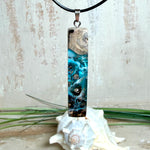 Rechteckiger Stabanhänger aus Maserholz und glasklarem Epoxidharz in tiefen Blau- und Türkistönen mit organischer Lagunen/Riff-Optik. Hochglanzpoliert, ca. 5,3 × 1,2 cm. Der Anhänger hängt an einer schwarzen Lederkette mit Edelstahl-Karabiner und 5-cm-Verlängerung (45 + 5 cm); die Anhängerschlaufe ist ebenfalls aus Edelstahl. Handgefertigtes Unikat, Lieferung in edler Schmuckbox – maritimer Look.