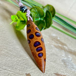 Handgefertigter Anhänger aus einem australischen Banksiazapfen mit eingelassenem metallic-violettem Harz. Der ovale, tropfenförmige Anhänger ist ca. 5,5 cm lang und wird mit einem verstellbaren Baumwollband geliefert. Jedes Stück zeigt die einzigartige natürliche Struktur des Zapfens und schimmert in satten Lilatönen – ein stilvoller Schmuck mit organischem Charakter.