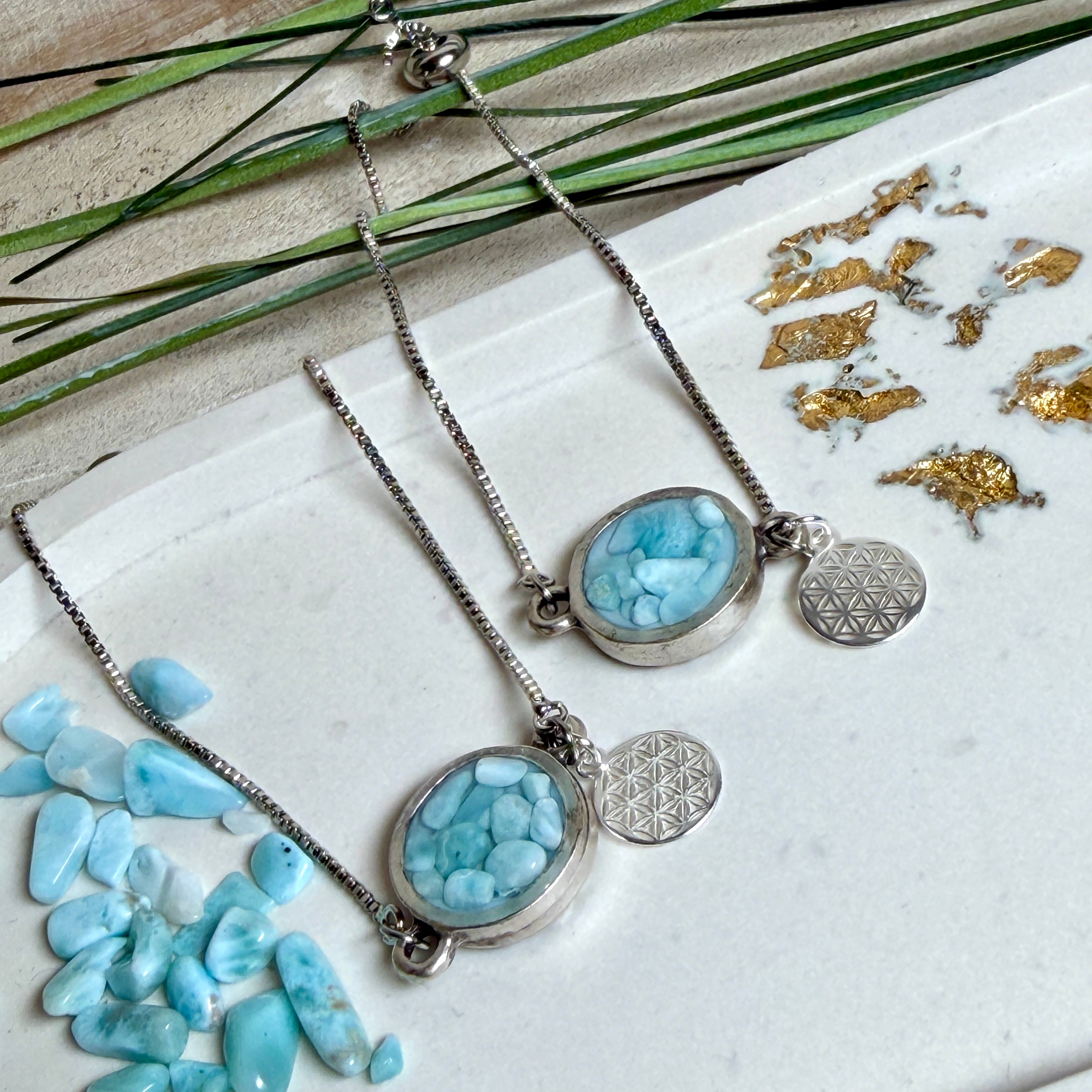 Verstellbares Edelstahlarmband mit Zirkoniasteinchen und Schiebeverschluss und rundem Anhänger (ca. 2,5 × 1,7 cm) mit echten Larimar-Edelstein-Splittern in Harz. Zusätzlich ein Charm „Blume des Lebens“ aus 925 Silber. Handgefertigtes Unikat in Türkis- und Meeresblautönen – Schmuckstück in Nahaufnahme bzw. als Tragebild am Handgelenk.