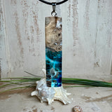 Rechteckiger Stabanhänger aus Maserholz und glasklarem Epoxidharz in tiefen Blau- und Türkistönen mit organischer Lagunen/Riff-Optik. Hochglanzpoliert, ca. 5,3 × 1,2 cm. Der Anhänger hängt an einer schwarzen Lederkette mit Edelstahl-Karabiner und 5-cm-Verlängerung (45 + 5 cm); die Anhängerschlaufe ist ebenfalls aus Edelstahl. Handgefertigtes Unikat, Lieferung in edler Schmuckbox – maritimer Look.