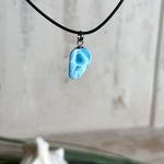 Larimar Freeform-Anhänger auf schwarzer Lederkette. Beidseitig tragbarer Trommelstein, ca. 2 x 1,8 cm groß, mit Silberöse. Der seltene, kostbare Larimar zeigt lebendige Blau- und Weißnuancen wie karibisches Lagunenwasser. Lederkette 45 cm mit Edelstahl-Karabiner und 5 cm Edelstahl-Verlängerung, fotografiert vor einer großen Muschel im maritimen Stil.