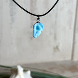 Larimar Freeform-Anhänger auf schwarzer Lederkette. Beidseitig tragbarer Trommelstein, ca. 2 x 1,8 cm groß, mit Silberöse. Der seltene, kostbare Larimar zeigt lebendige Blau- und Weißnuancen wie karibisches Lagunenwasser. Lederkette 45 cm mit Edelstahl-Karabiner und 5 cm Edelstahl-Verlängerung, fotografiert vor einer großen Muschel im maritimen Stil.