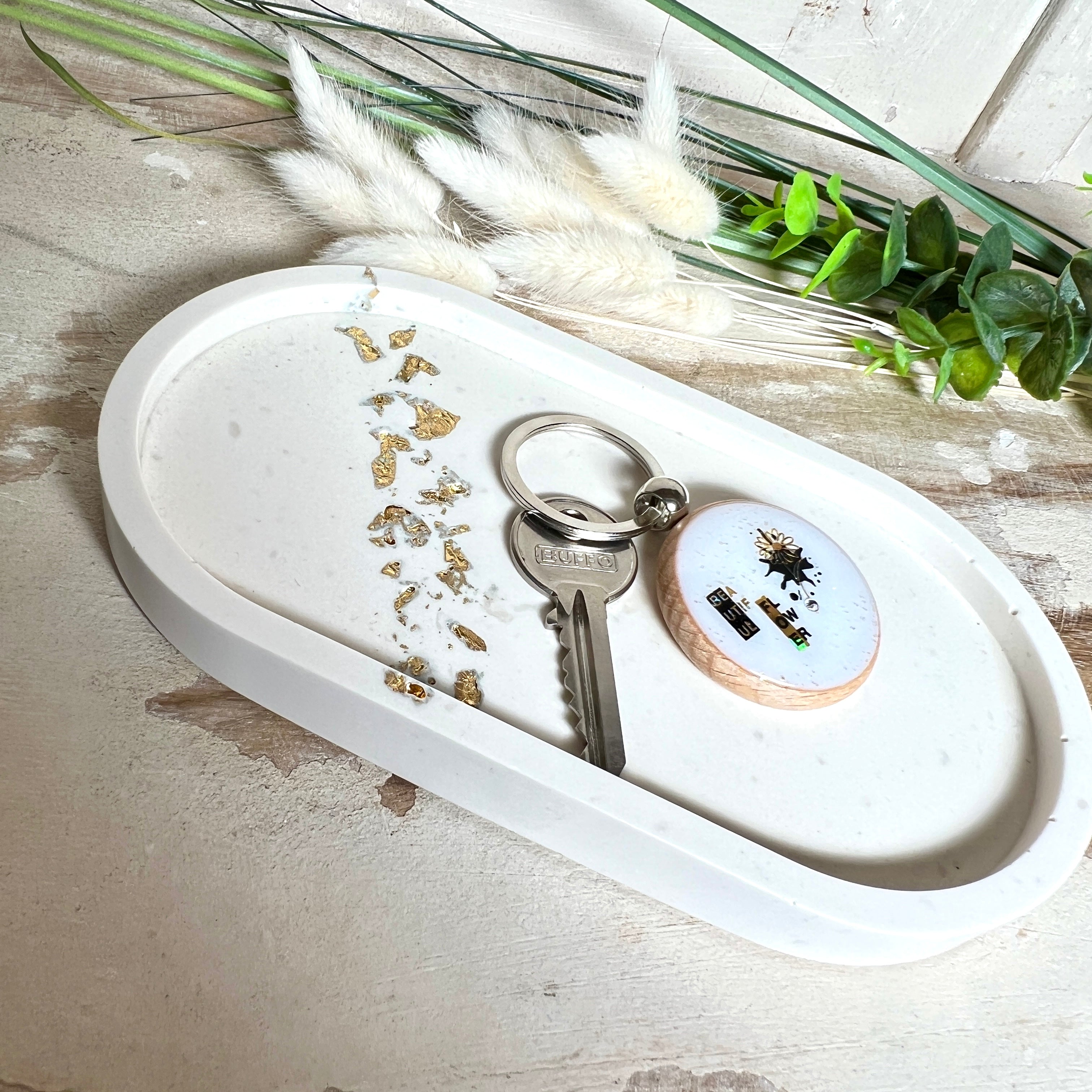 Handgefertigtes ovales Schmucktablett aus Jesmonite mit schimmernden Blattgold-Akzenten. Die Rückseite ist mit rutschfesten Korkpunkten ausgestattet. Ideal zur stilvollen Aufbewahrung von Schmuck, Schlüsseln oder kleinen Accessoires auf Kommoden und Nachttischen. Helles Design mit natürlicher Oberfläche und modernem Look. Unikat in feiner Handarbeit gefertigt – ohne Deko geliefert.