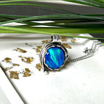 Handgemachter Anhänger mit rundem Cabochon aus Epoxidharz und schimmerndem synthetischem Opal. Die Farben changieren je nach Lichteinfall in Blau, Türkis und Grün. Der Opal sitzt in einer antik versilberten Fassung aus Messing (blei- & nickelfrei). Geliefert mit einer rhodinierten 925 Silberkette (45 cm). Jedes Stück ein Einzelstück – perfekt als stilvolles Geschenk oder persönlicher Glücksbringer.
