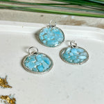 Runder Anhänger „Atlantis Dream“ aus antik-silberfarbener Zinklegierung, gefüllt mit kleinen Larimar-Edelsteinchen und klarem Resin. Die Larimarsteine zeigen sanfte Blau- und Weißtöne, die an das Meer erinnern. Durchmesser ca. 22 mm, handgefertigtes Unikat, fotografiert auf maritimem Untergrund mit kleinen Larimarsteinen und Muscheln zur Größenorientierung.