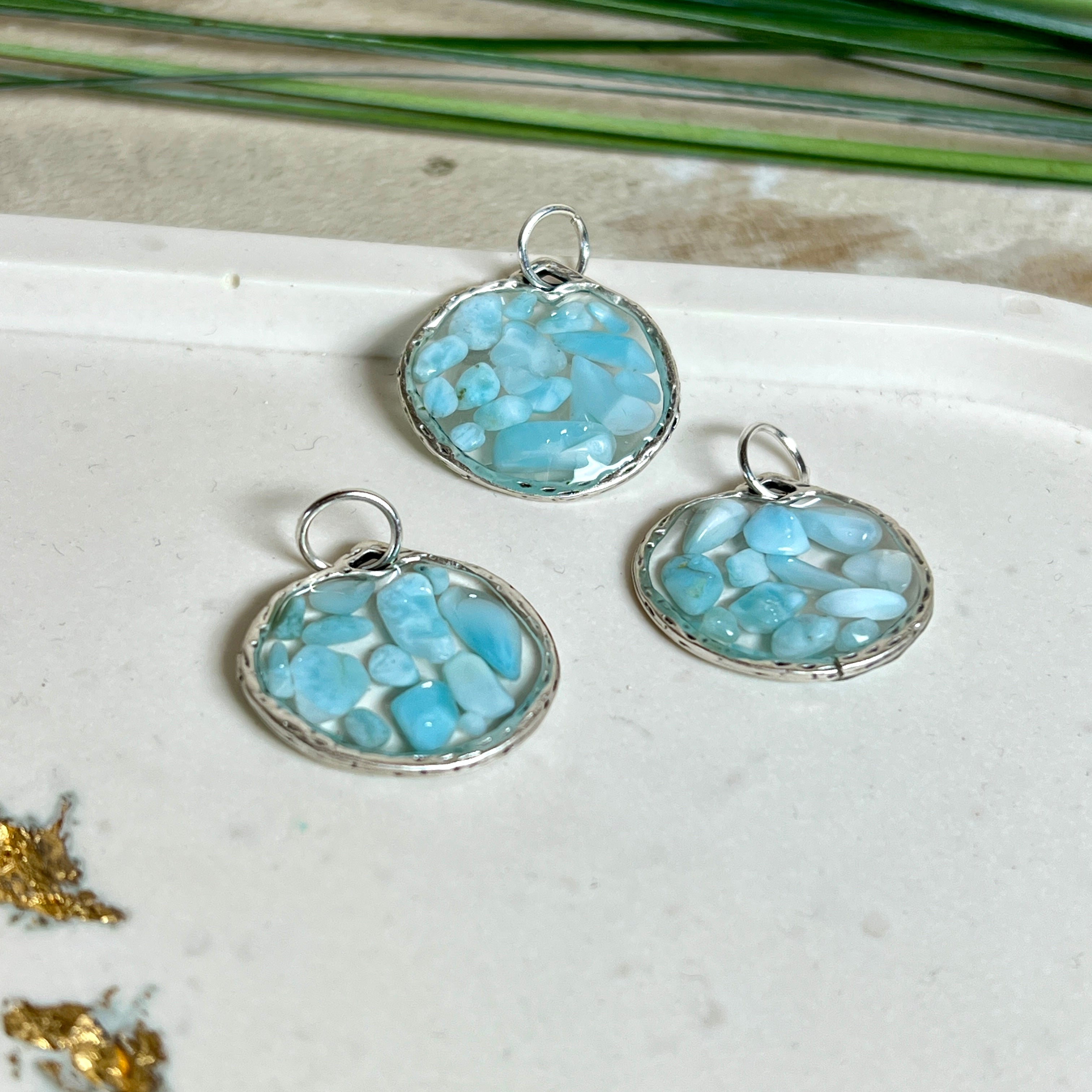 Runder Anhänger „Atlantis Dream“ aus antik-silberfarbener Zinklegierung, gefüllt mit kleinen Larimar-Edelsteinchen und klarem Resin. Die Larimarsteine zeigen sanfte Blau- und Weißtöne, die an das Meer erinnern. Durchmesser ca. 22 mm, handgefertigtes Unikat, fotografiert auf maritimem Untergrund mit kleinen Larimarsteinen und Muscheln zur Größenorientierung.