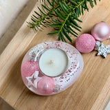Rosa Teelichthalter aus klarem Epoxidharz, ca. 8 × 6,5 cm, Höhe ca. 4 cm. Im Harz sind kleine rosa Weihnachtskugeln, ein Stern, eine Schneeflocke und rosa Granulat eingegossen. Ovaler Halter für ein Teelicht, Unterseite mit vier runden Korkpads als Möbelschoner. Fotografiert auf Holz mit Tannenzweig und rosa Kugeln als weihnachtlicher Dekohintergrund.