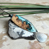 Handgefertigter, länglicher Surfbrett-Anhänger aus naturbelassenem Maserholz und transparentem Harz in Lagunen-Türkis, an schwarzem Lederband mit Knoten – maritimer Unikatschmuck für Damen und Herren, dekorativ mit Stein und Muschel inszeniert.