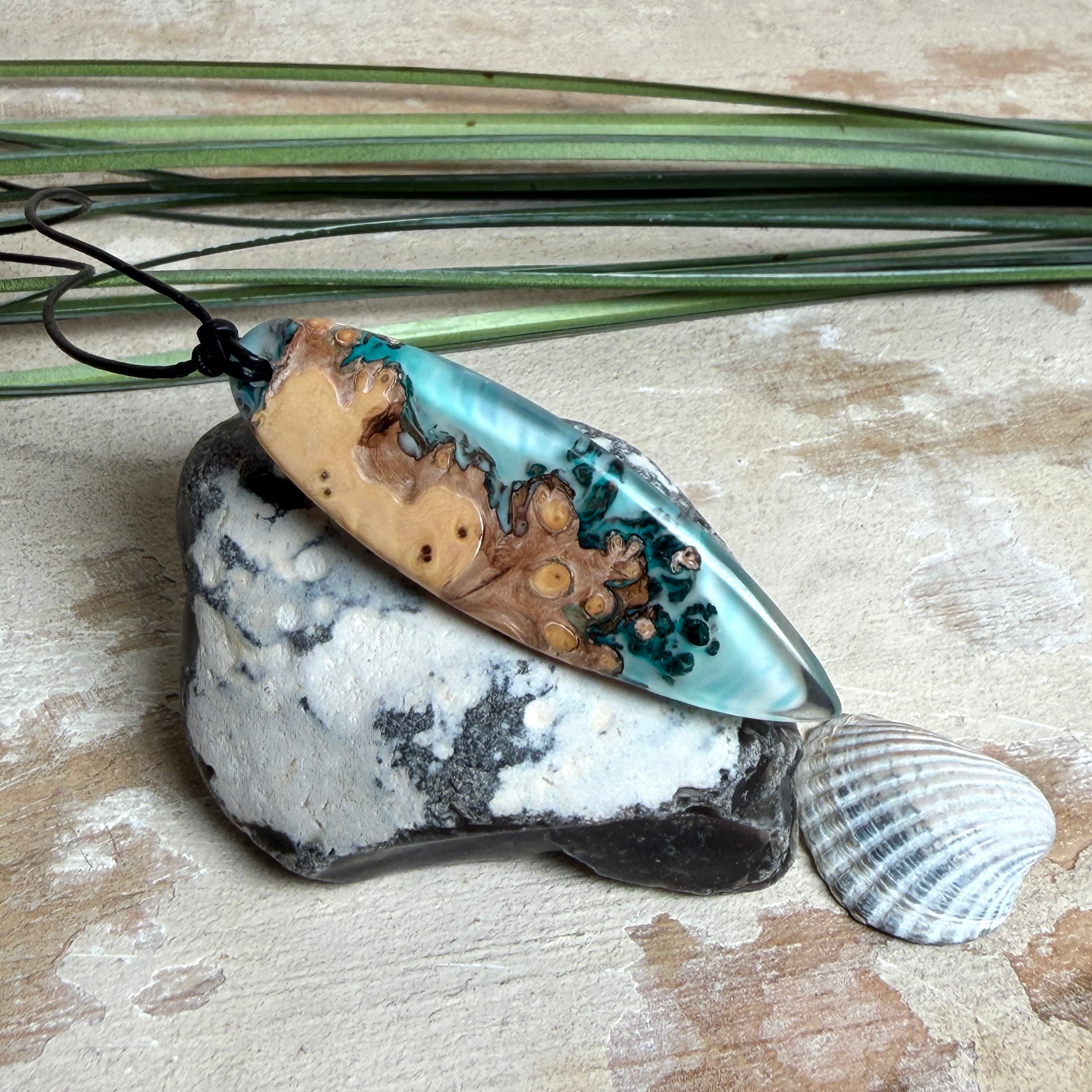 Handgefertigter, länglicher Surfbrett-Anhänger aus naturbelassenem Maserholz und transparentem Harz in Lagunen-Türkis, an schwarzem Lederband mit Knoten – maritimer Unikatschmuck für Damen und Herren, dekorativ mit Stein und Muschel inszeniert.