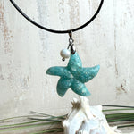 Seestern-Anhänger aus transparentem Harz mit echtem Meersand ( leicht eingefärbt), in blautürkis Strand-Look, mit kleiner Süßwasserperle als Anhänger-Detail. Edelstahlöse, schwarze Lederkette mit Edelstahl-Karabiner und Verlängerung (45 cm + 5 cm). Anhänger ca. 3,5 × 3,5 cm. Handgefertigtes Unikat; vereinzelt kleine Luftbläschen möglich, typisch für Handarbeit.