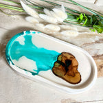 Handgefertigtes Schmucktablett aus Jesmonite mit eingebettetem Olivenholz und türkisfarbenem Resin. Die glänzende Oberfläche erinnert an Wasserstrukturen, während das Naturholz Tiefe und Wärme verleiht. Die Rückseite ist mit Korkpunkten versehen und schützt empfindliche Oberflächen. Perfekt als Ablage für Schmuck, Brille, Schlüssel oder Deko jedes Stück ein Unikat.