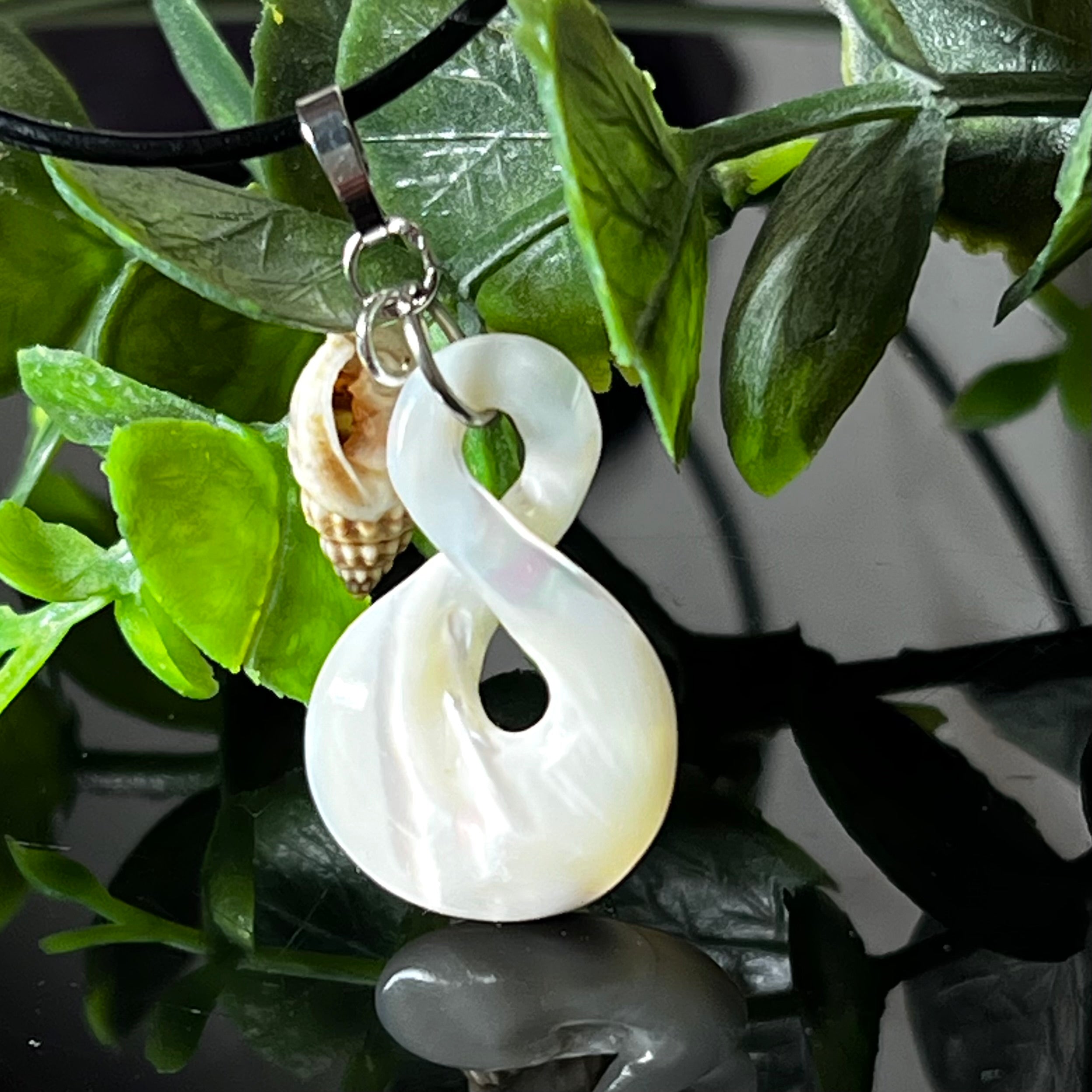 Detailaufnahme eines handgefertigten Anhängers aus echtem Perlmutt in Schlaufenform, kombiniert mit einer kleinen echten Muschel als Charm-Anhänger. Der Schmuck hängt an einer feinen schwarzen Lederschnur mit Edelstahlverschluss und symbolisiert Harmonie und Unendlichkeit. Ideal für Meeresliebhaberinnen und Fans von Naturmaterialien.