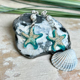 Handgefertigte Seestern-Ohrringe aus Epoxidharz und Sand mit Süßwasserperle, an 925 Sterling Silber Ohrsteckern; versilberte Anhängerschlaufe. Je Seestern ca. 2,6 × 2,5 cm. Leichte, maritime Dangle-Ohrringe im Strand-Look – ideal als Geschenk für Meeresliebhaberinnen.
