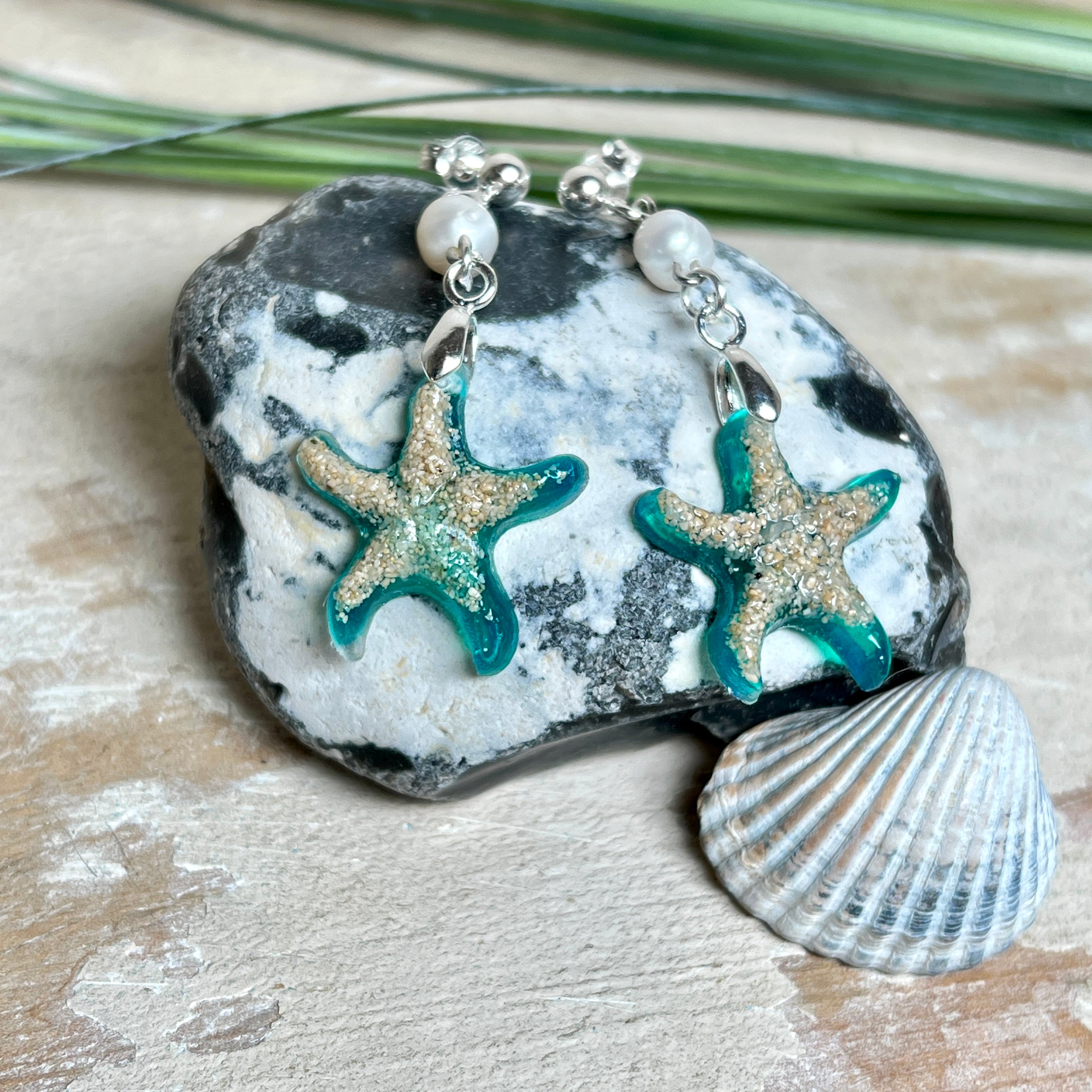 Handgefertigte Seestern-Ohrringe aus Epoxidharz und Sand mit Süßwasserperle, an 925 Sterling Silber Ohrsteckern; versilberte Anhängerschlaufe. Je Seestern ca. 2,6 × 2,5 cm. Leichte, maritime Dangle-Ohrringe im Strand-Look – ideal als Geschenk für Meeresliebhaberinnen.