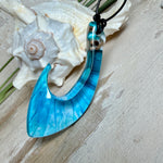 Handgefertigter Haken-Anhänger im Hei-Matau-Stil aus Maserholz und klarem Harz mit Blau-, Türkis- und Weißverlauf, hochglanzpoliert. Oben sichtbares Holz im Harzbereich, getragen an verstellbarem schwarzem Lederband mit Schiebeknoten und Zierperlen. Anhänger ca. 7 × 3 cm, fotografiert mit Muscheln im Hintergrund.