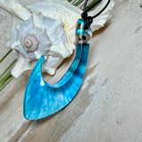 Handgefertigter Haken-Anhänger im Hei-Matau-Stil aus Maserholz und klarem Harz mit Blau-, Türkis- und Weißverlauf, hochglanzpoliert. Oben sichtbares Holz im Harzbereich, getragen an verstellbarem schwarzem Lederband mit Schiebeknoten und Zierperlen. Anhänger ca. 7 × 3 cm, fotografiert mit Muscheln im Hintergrund.