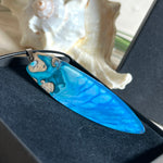 Surfbrett-Anhänger aus Maserholz und blau-weißem Epoxidharz, hochglanzpoliert, ca. 6,5 × 2 cm. An schwarzer Lederkette mit Edelstahl-Karabiner und 5-cm-Verlängerung; Edelstahl-Öse. Im Tageslicht lagunenblau, unter Schwarzlicht fluoreszierend – ideal als Party-/Festival-Schmuck. Handgefertigtes Unikat, Lieferung in edler Schmuckbox.
