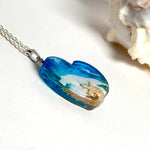 Ein herzförmiger Anhänger mit einer ganzen Ozeanwelt darin: Welle, Sand und Seestern in leuchtendem Blau und Türkis – handgegossen, einzigartig, unvergesslich. Mit versilberter Kette (46 cm) und Edelstahl-Karabiner.