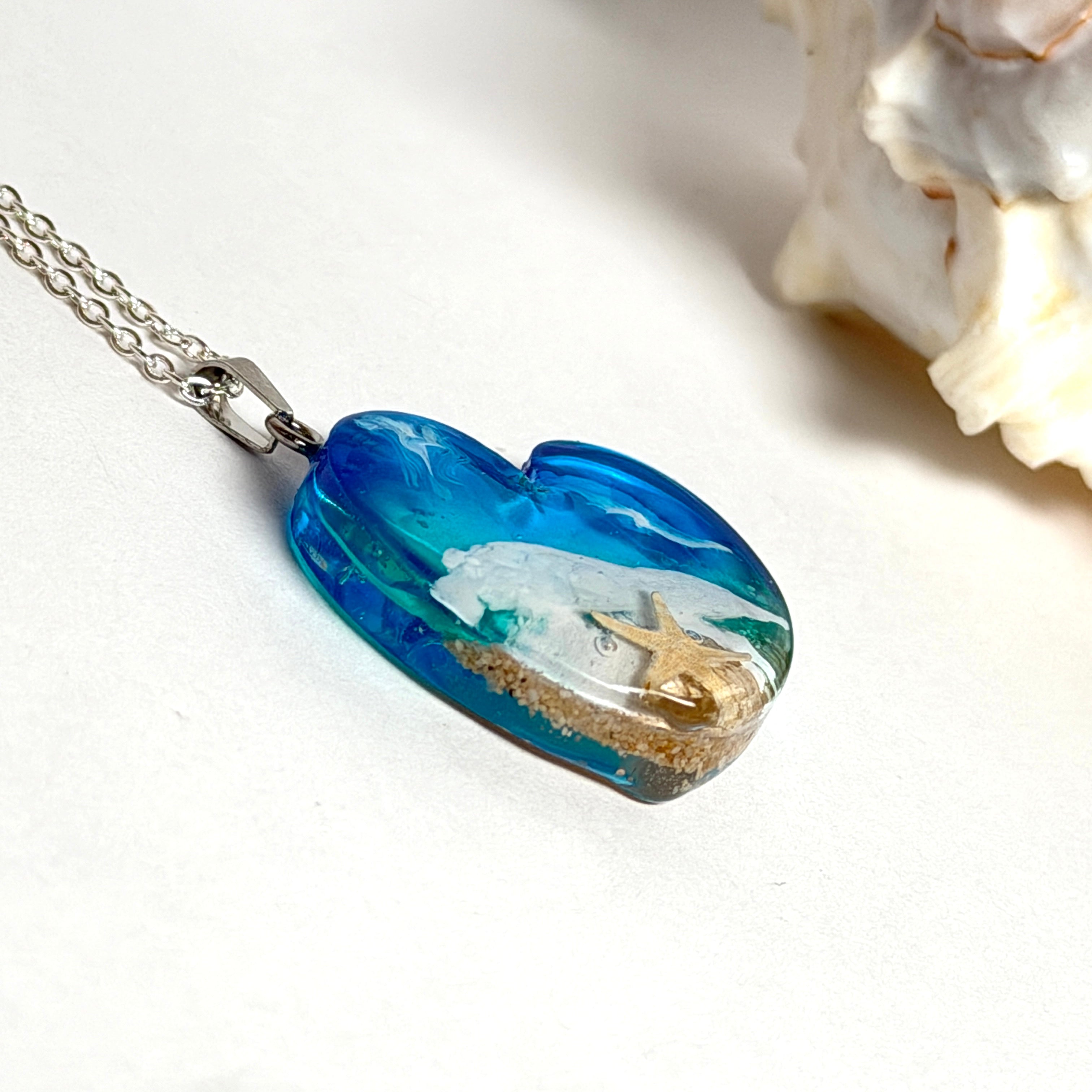Ein herzförmiger Anhänger mit einer ganzen Ozeanwelt darin: Welle, Sand und Seestern in leuchtendem Blau und Türkis – handgegossen, einzigartig, unvergesslich. Mit versilberter Kette (46 cm) und Edelstahl-Karabiner.