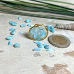 Runder Anhänger „Golden Atlantis Dream“ mit kleinen Larimar-Edelsteinchen in klarem Resin, gefasst in einer 24K vergoldeten Zinklegierung in Shiny Gold. Die Larimarsteine zeigen sanfte Blau- und Weißtöne wie karibische Wellen. Durchmesser ca. 22 mm, handgefertigtes Unikat, fotografiert mit Larimarsteinchen, Muscheln und einer 2-Euro-Münze zur Größenorientierung auf maritimem Untergrund.