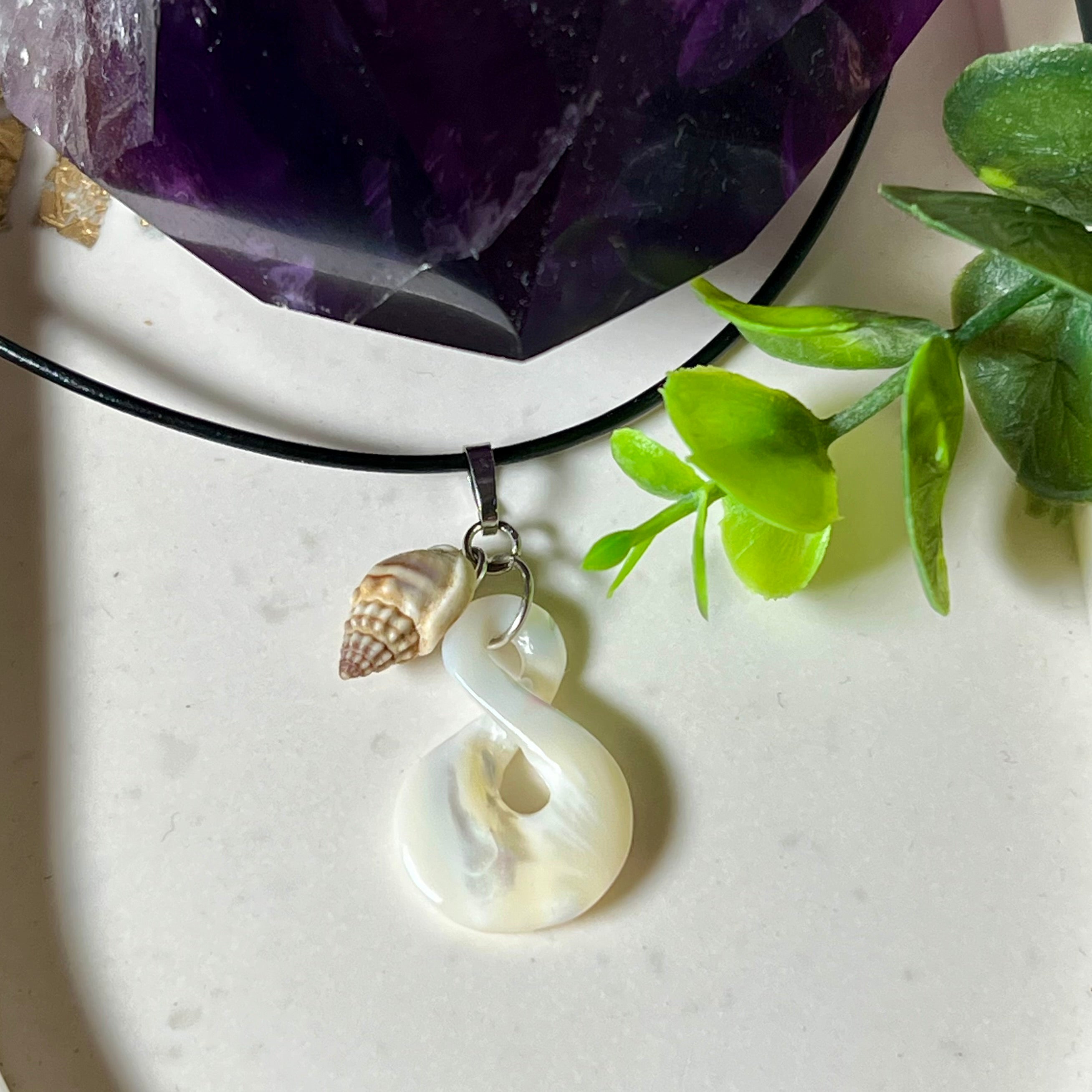 Detailaufnahme eines handgefertigten Anhängers aus echtem Perlmutt in Schlaufenform, kombiniert mit einer kleinen echten Muschel als Charm-Anhänger. Der Schmuck hängt an einer feinen schwarzen Lederschnur mit Edelstahlverschluss und symbolisiert Harmonie und Unendlichkeit. Ideal für Meeresliebhaberinnen und Fans von Naturmaterialien.