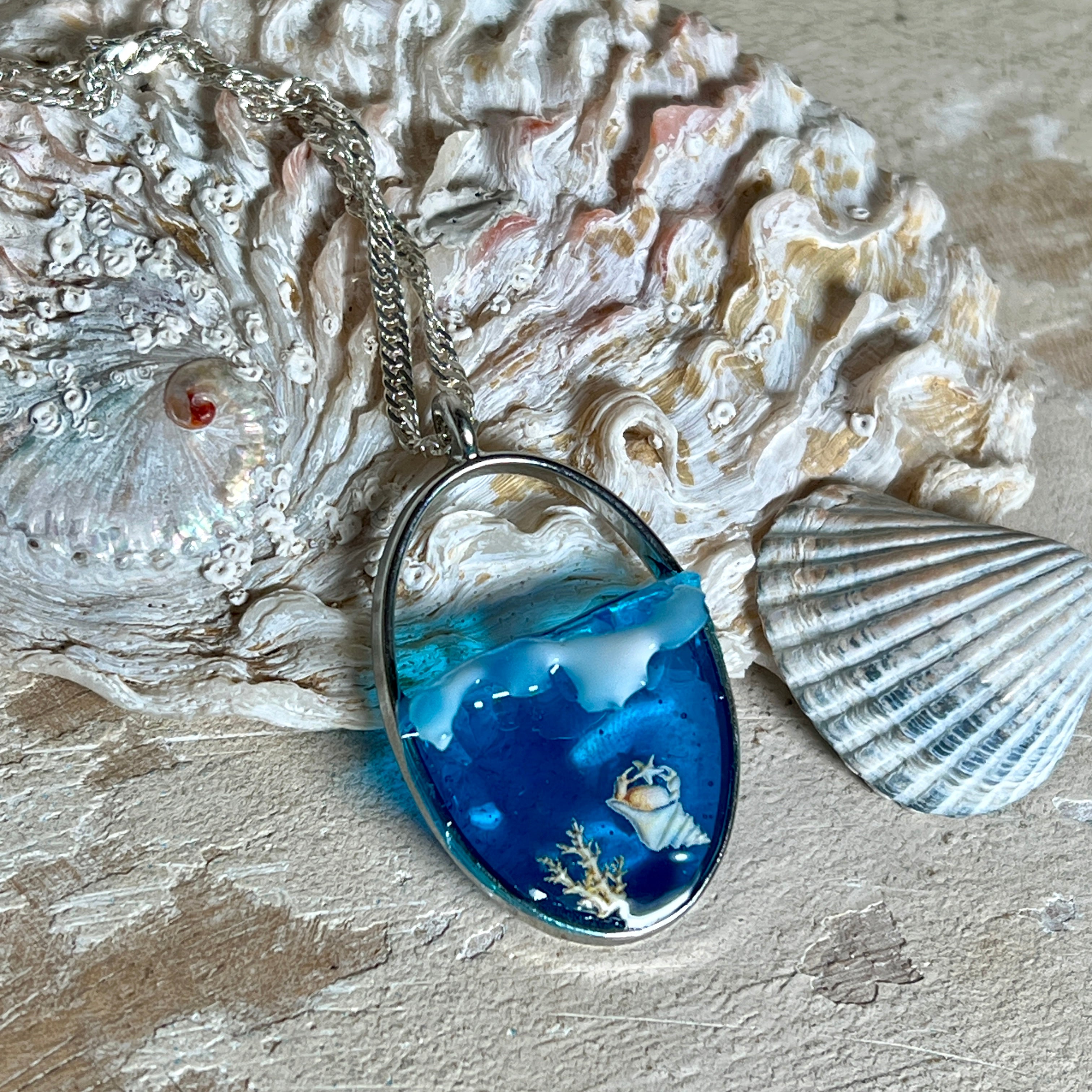 „Riffschatz“ – ovaler Harz-Anhänger mit plastischer 3D-Welle in Blau/Türkis, weißer Schaumkrone und Muschel-/Korallen-Stickern am Grund. Die Sticker sind unter klarem Harz versiegelt. Silber-Legierung, ca. 3,9 × 2,3 cm, an einer ca. 56 cm langen versilberten Kette. Handgefertigt, hochglänzend, jedes Stück ein Unikat.