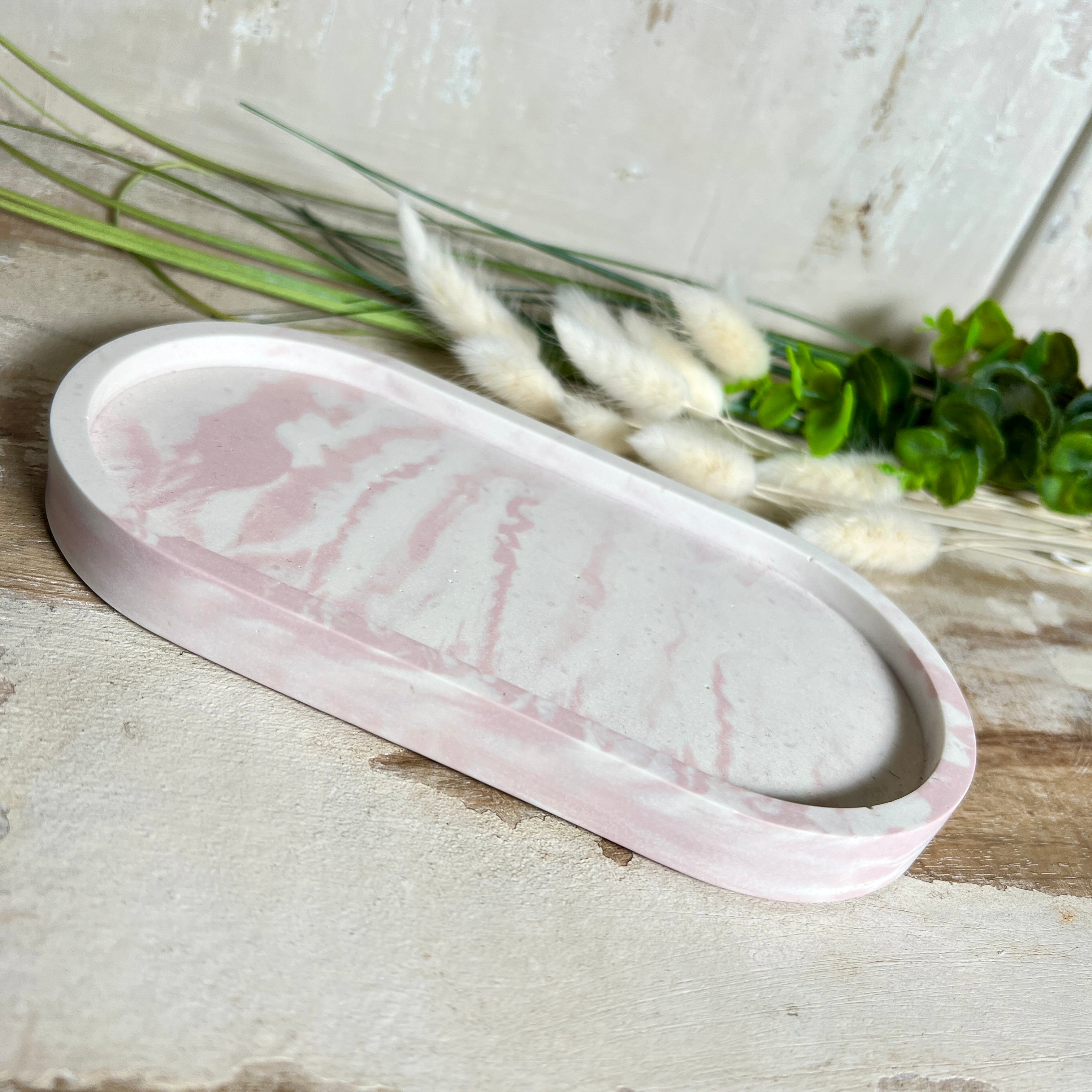 Schmucktablett aus Jesmonite in zartem Rosa-Weiß-Marmor. Handgegossenes, ovales Deko-Tablett mit glatter Oberfläche und Korkfüßen. Perfekt als Ablage für Schmuck, Schlüssel, Brille oder Dekoobjekte. Jedes Stück ein liebevoll gefertigtes Unikat in der Größe 17,5 × 9,5 × 1,7 cm.
