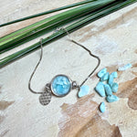 Verstellbares Edelstahlarmband mit Zirkoniasteinchen und Schiebeverschluss und rundem Anhänger (ca. 2,5 × 1,7 cm) mit echten Larimar-Edelstein-Splittern in Harz. Zusätzlich ein Charm „Blume des Lebens“ aus 925 Silber. Handgefertigtes Unikat in Türkis- und Meeresblautönen – Schmuckstück in Nahaufnahme bzw. als Tragebild am Handgelenk.