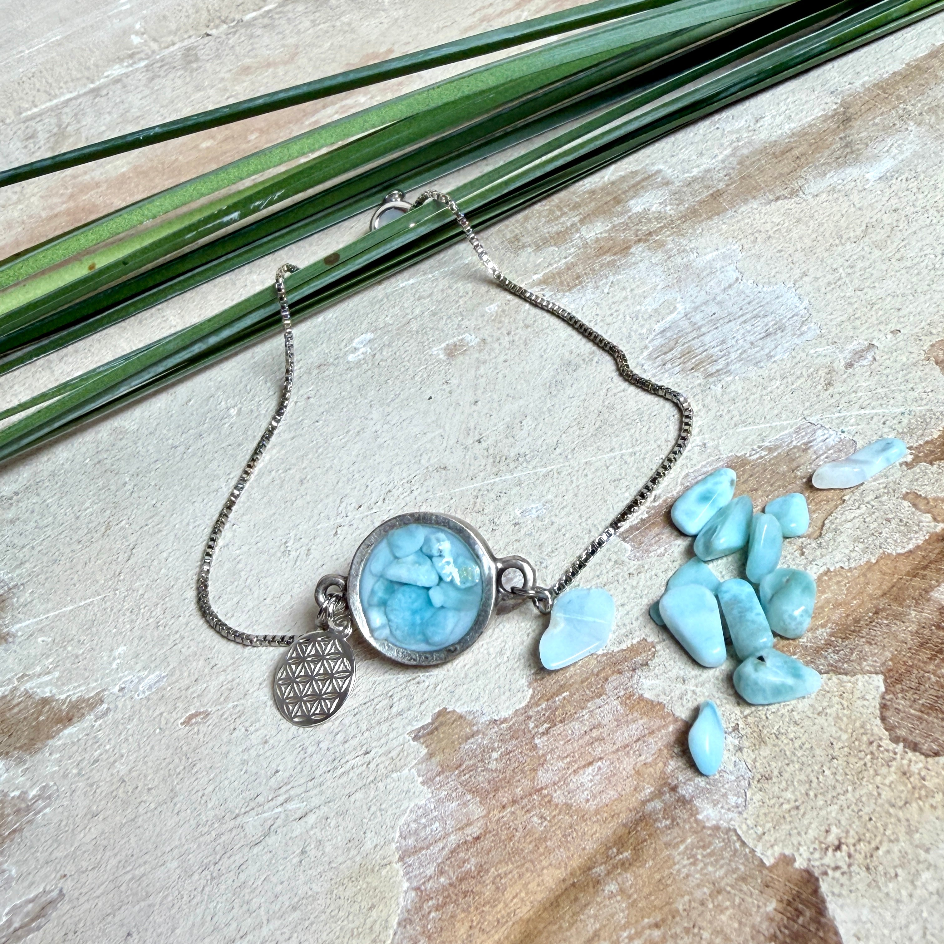 Verstellbares Edelstahlarmband mit Zirkoniasteinchen und Schiebeverschluss und rundem Anhänger (ca. 2,5 × 1,7 cm) mit echten Larimar-Edelstein-Splittern in Harz. Zusätzlich ein Charm „Blume des Lebens“ aus 925 Silber. Handgefertigtes Unikat in Türkis- und Meeresblautönen – Schmuckstück in Nahaufnahme bzw. als Tragebild am Handgelenk.