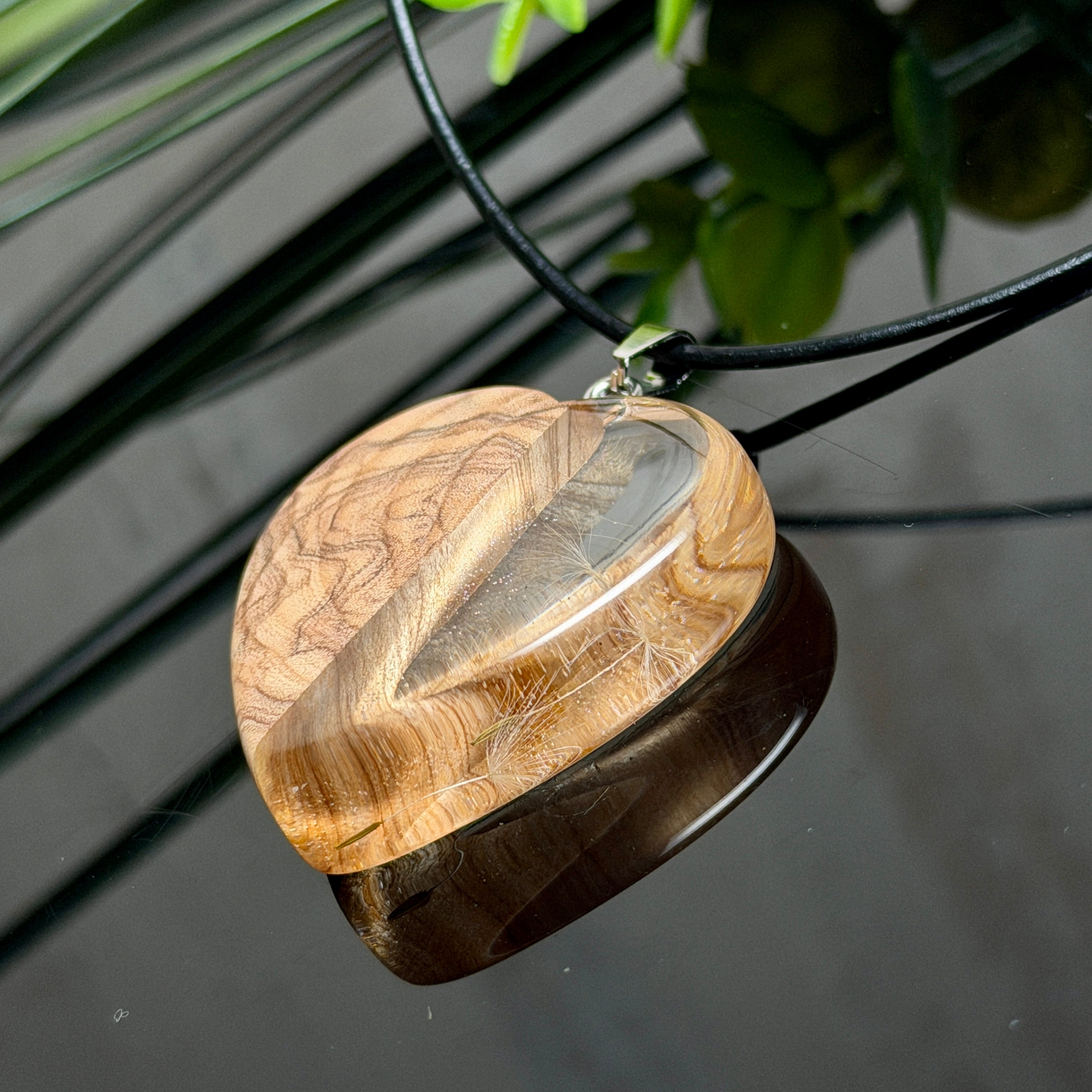 Pusteblumen Herzanhänger aus Olivenholz und glasklarem Epoxidharz, echte Löwenzahnsamen im Harz schwebend, schwarze Lederkette, handgefertigter Naturschmuck mit Spiegelung, dunkler Hintergrund