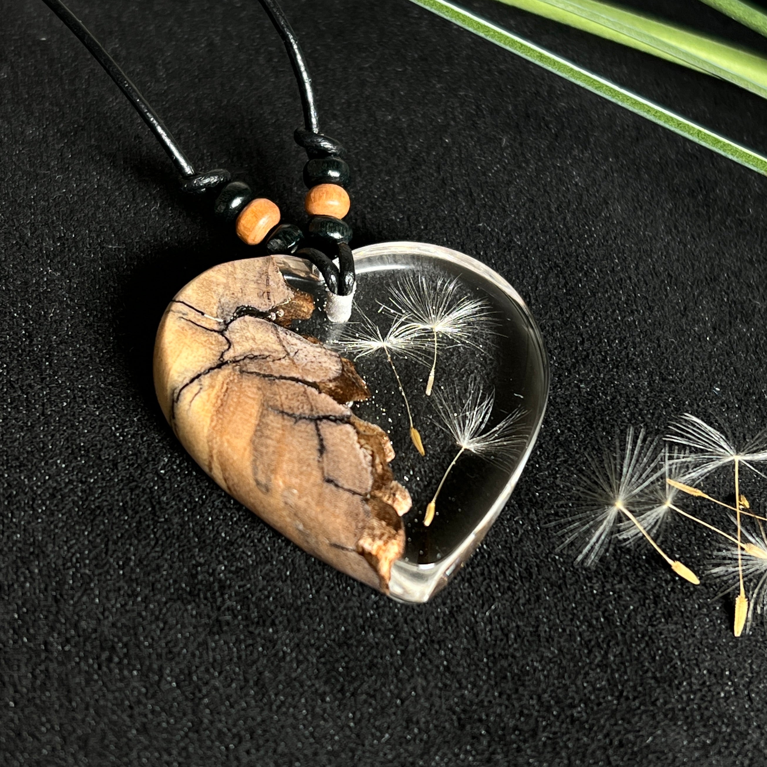 „Handgefertigter Herzanhänger aus Olivenholz und klarem Harz mit echter Pusteblume. Der Anhänger (ca. 3,5 × 3,5 cm) hängt an einem verstellbaren Lederband (23–43 cm) mit Holzperlen. Symbol für Wünsche und Glück, sorgfältig in Handarbeit gegossen, gesägt, geschliffen und poliert. Ein einzigartiger, naturinspirierter Anhänger – ideal als Geschenk oder persönlicher Talisman.“