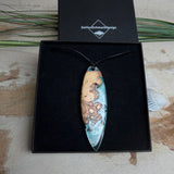 Handgefertigter, länglicher Surfbrett-Anhänger aus naturbelassenem Maserholz und transparentem Harz in Lagunen-Türkis, an schwarzem Lederband mit Knoten – maritimer Unikatschmuck für Damen und Herren, dekorativ mit Stein und Muschel inszeniert.