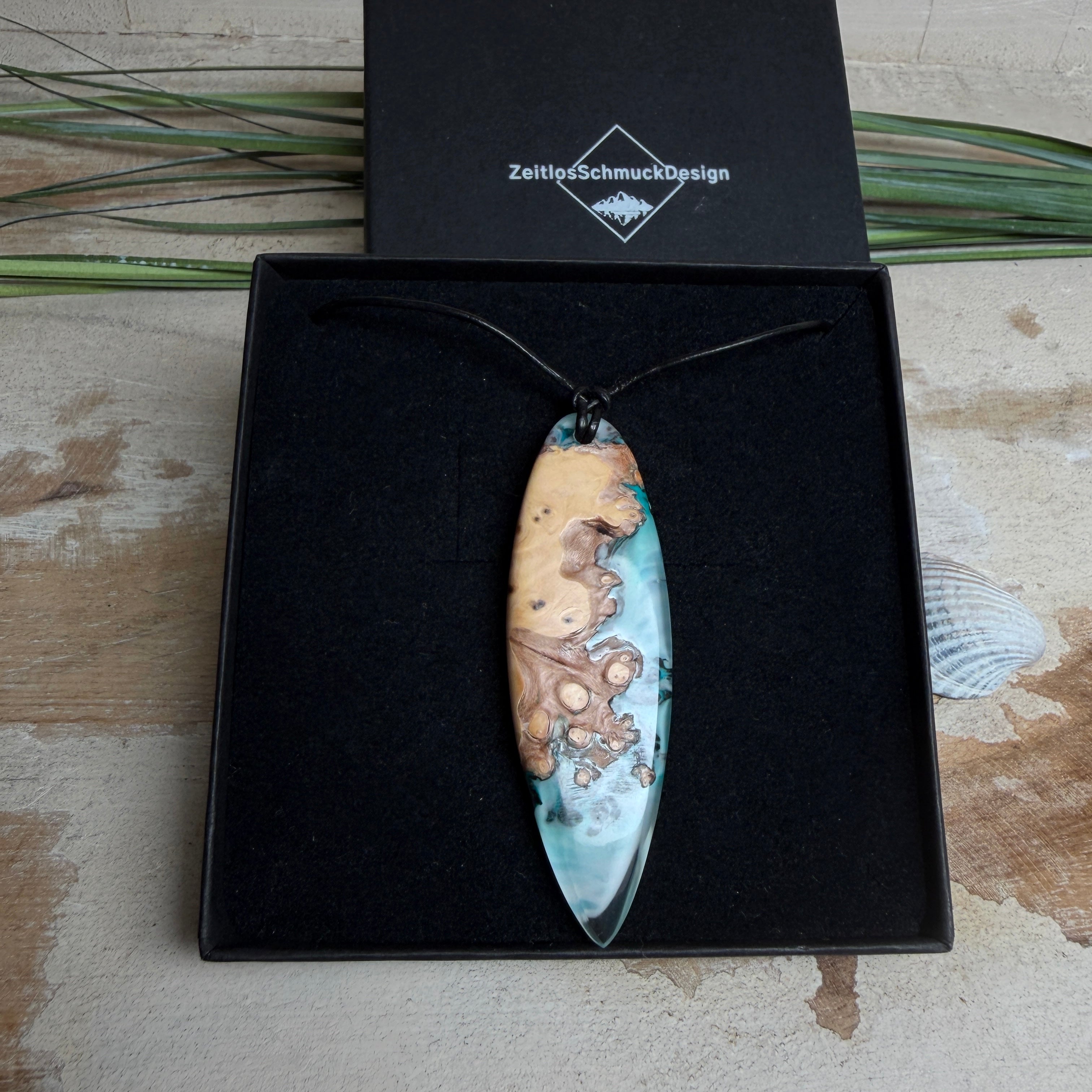 Handgefertigter, länglicher Surfbrett-Anhänger aus naturbelassenem Maserholz und transparentem Harz in Lagunen-Türkis, an schwarzem Lederband mit Knoten – maritimer Unikatschmuck für Damen und Herren, dekorativ mit Stein und Muschel inszeniert.