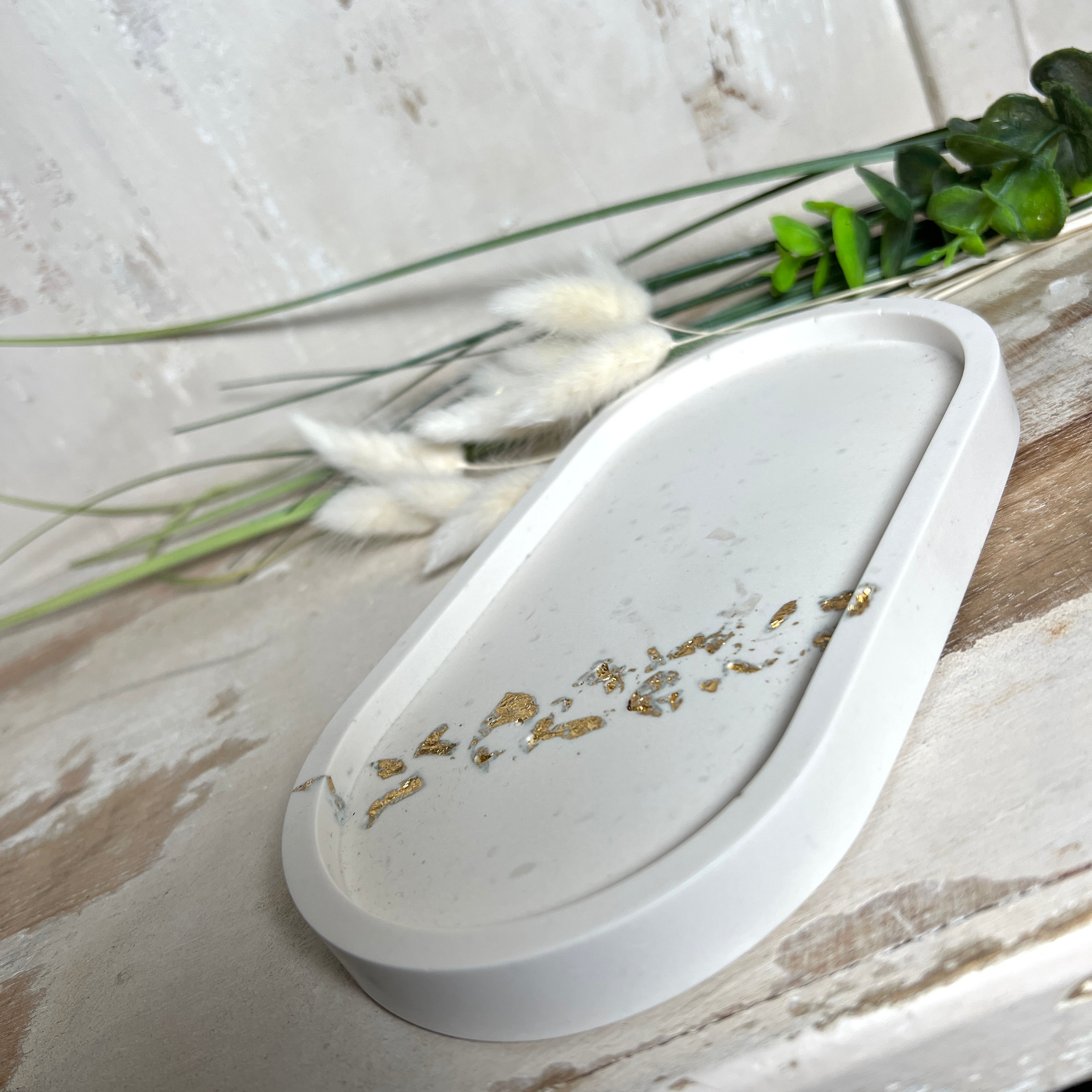 Handgefertigtes ovales Schmucktablett aus Jesmonite mit schimmernden Blattgold-Akzenten. Die Rückseite ist mit rutschfesten Korkpunkten ausgestattet. Ideal zur stilvollen Aufbewahrung von Schmuck, Schlüsseln oder kleinen Accessoires auf Kommoden und Nachttischen. Helles Design mit natürlicher Oberfläche und modernem Look. Unikat in feiner Handarbeit gefertigt – ohne Deko geliefert.