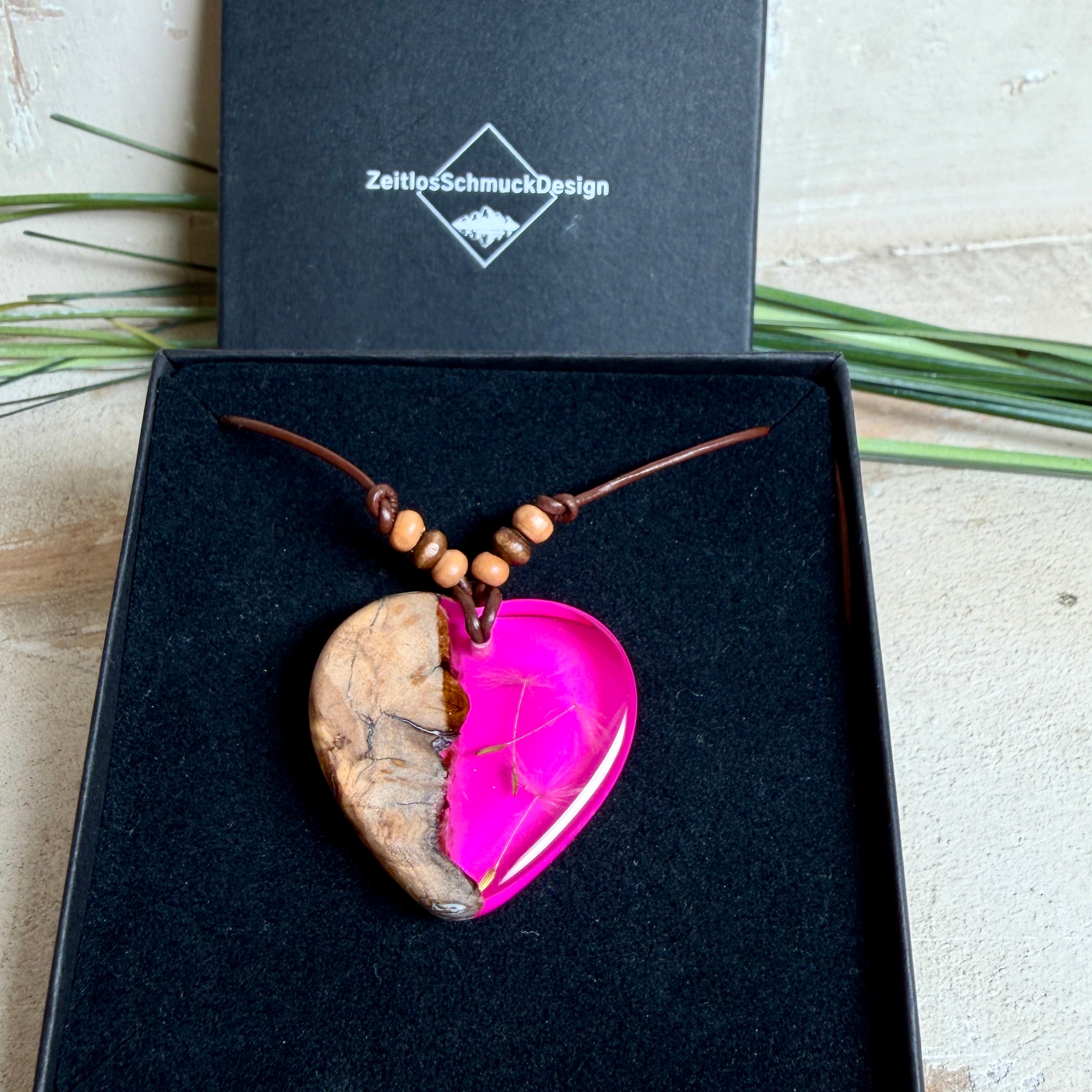 Handgefertigter Herzanhänger aus Olivenholz und leuchtend pinkem Epoxidharz mit eingeschlossenen Pusteblumensamen. Verstellbares braunes Lederband mit Holzperlen, Anhänger ca. 3,5 × 3,3 cm groß. Das Harz leuchtet dezent unter Schwarzlicht. Ein einzigartiges Unikat von ZeitlosSchmuckDesign, inspiriert von Kindheitsglück und Pusteblumen.