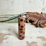 Handgefertigter Halsanhänger aus echtem Banksiazapfen, veredelt mit rotem Harz in den natürlichen Kammern; ca. 5,5 × 1,6 cm. Mit Anhängerschlaufe aus Edelstahl an schwarzer Lederkette (45 cm + 5 cm Verlängerung) mit Edelstahl-Karabiner. Detailaufnahme bzw. Tragebild vor neutralem, natürlichem Hintergrund.