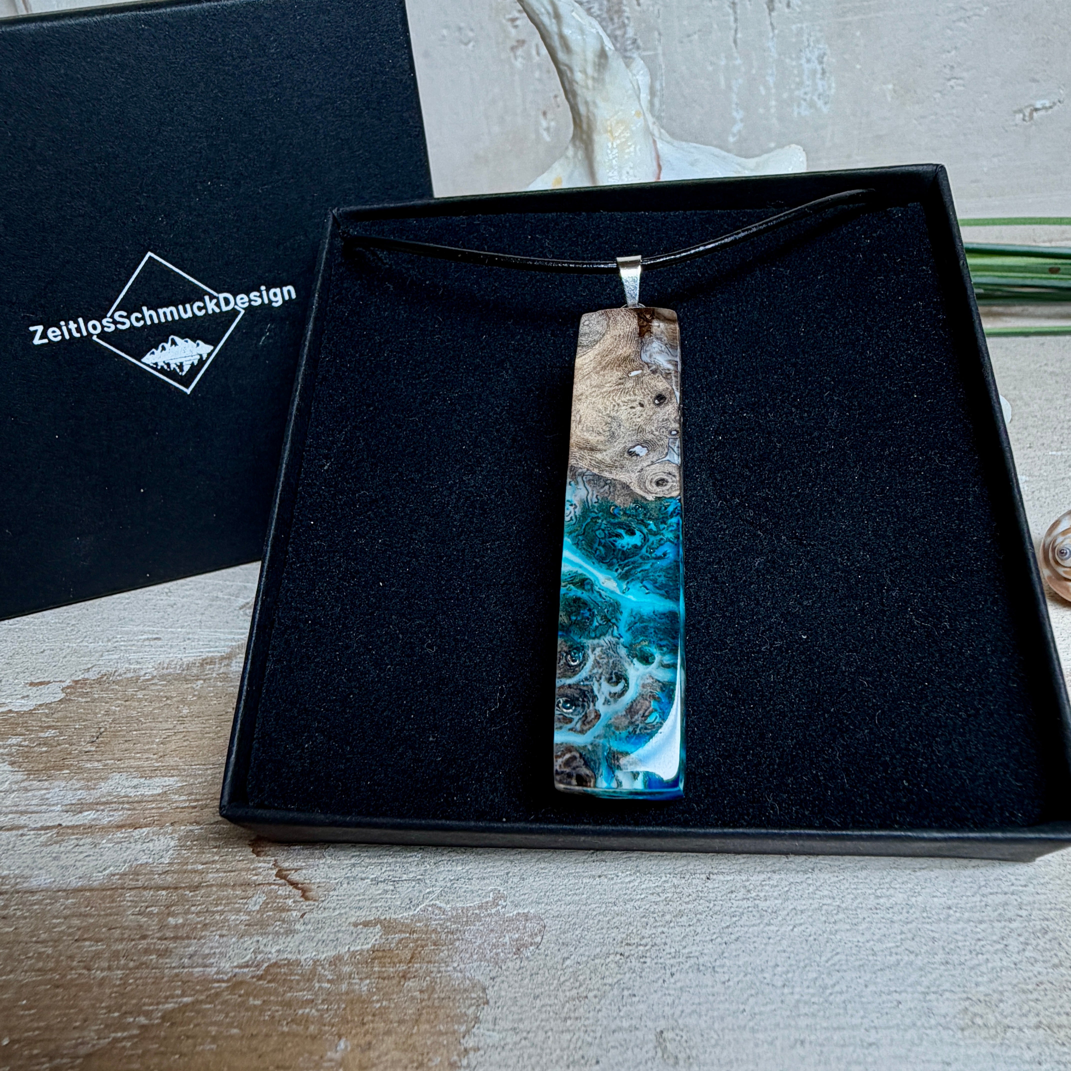 Rechteckiger Stabanhänger aus Maserholz und glasklarem Epoxidharz in tiefen Blau- und Türkistönen mit organischer Lagunen/Riff-Optik. Hochglanzpoliert, ca. 5,3 × 1,2 cm. Der Anhänger hängt an einer schwarzen Lederkette mit Edelstahl-Karabiner und 5-cm-Verlängerung (45 + 5 cm); die Anhängerschlaufe ist ebenfalls aus Edelstahl. Handgefertigtes Unikat, Lieferung in edler Schmuckbox – maritimer Look.