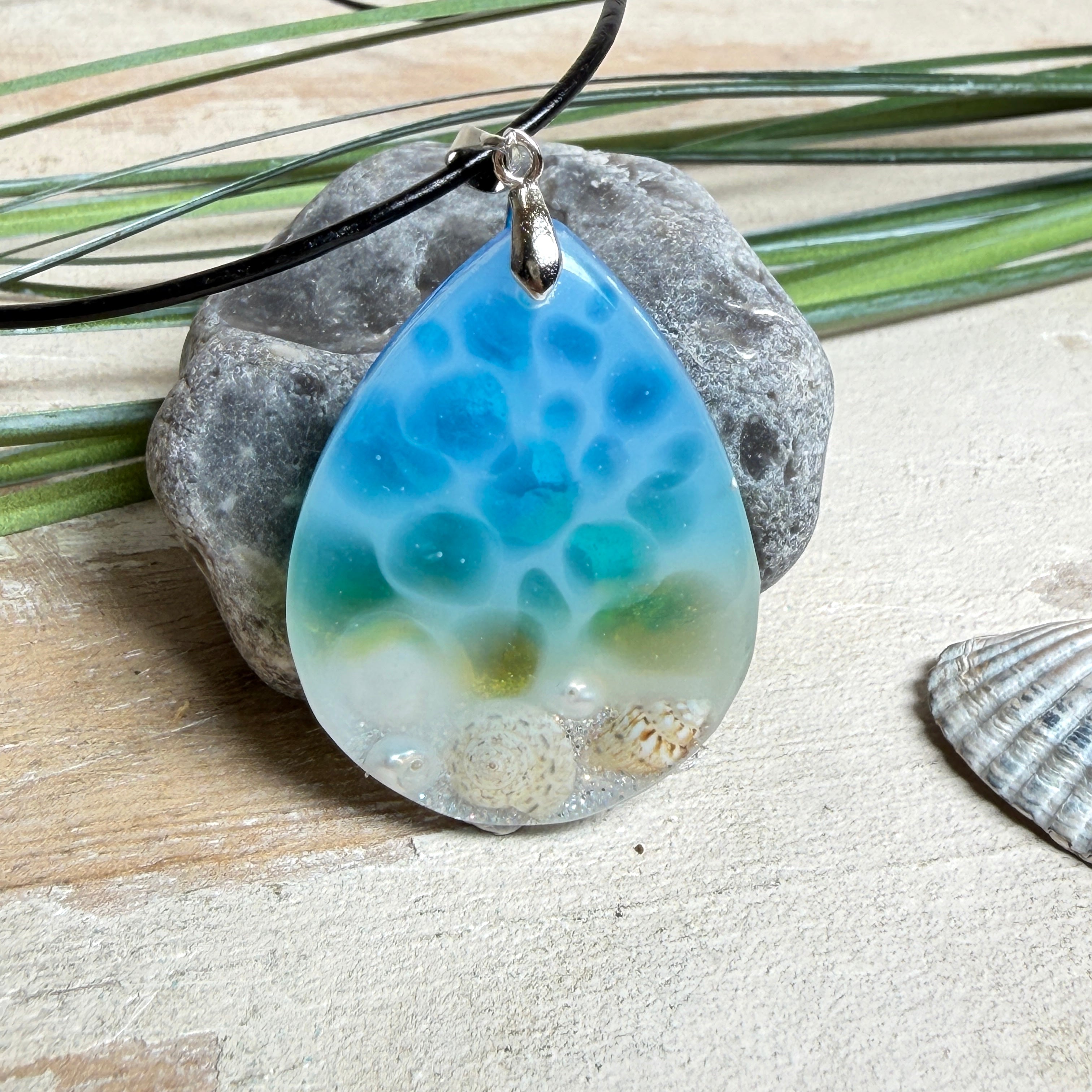 Tropfenförmiger Harzanhänger „Lagoon Drop“ in Blau-Grün-Weiß mit ca. 4 x 3 cm Größe. Im unteren Bereich sind kleine Muscheln, Sandstruktur und feine Süßwasserperlen eingegossen, sodass der Eindruck eines seichten Strandes im klaren Wasser entsteht. Der Anhänger hat eine Edelstahl-Schlaufe und hängt an einer schwarzen Lederkette mit Edelstahl-Karabiner und Verlängerung. Handgefertigter, maritime Unikat-Schmuck für Meeresliebhaberinnen.