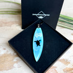 Heller Holzhintergrund: Surfbrett Anhänger aus hellblauem Epoxidharz mit schwarzer Schildkröten-Silhouette und weißem Welleneffekt, schwarze Lederkette, handgefertigter maritimer Naturschmuck.