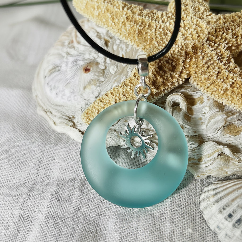 Handgefertigte Halskette mit Donut-Anhänger aus türkisfarbenem Seeglas, kombiniert mit einem filigranen Sonnen-Charm aus Edelstahl. Die Kette besteht aus einer schwarzen Lederschnur (1,5 mm) mit Karabiner und Verlängerungskette aus Edelstahl. Maritimes Schmuckstück für Meerliebhaberinnen und Sommerfreunde. Unikat mit stilvoller Ausstrahlung.