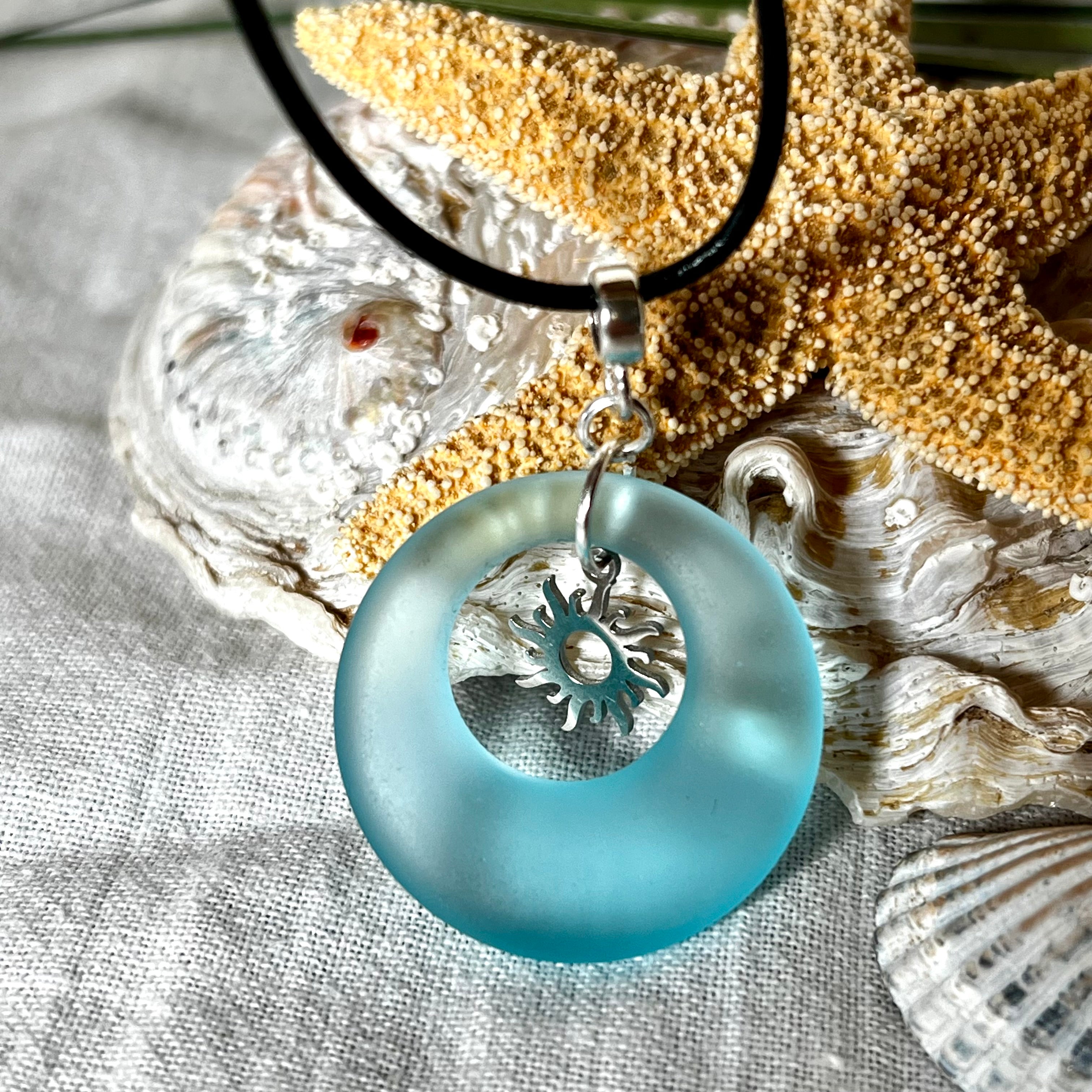 Handgefertigte Halskette mit Donut-Anhänger aus türkisfarbenem Seeglas, kombiniert mit einem filigranen Sonnen-Charm aus Edelstahl. Die Kette besteht aus einer schwarzen Lederschnur (1,5 mm) mit Karabiner und Verlängerungskette aus Edelstahl. Maritimes Schmuckstück für Meerliebhaberinnen und Sommerfreunde. Unikat mit stilvoller Ausstrahlung.